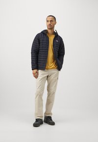 Fekete steppelt dzseki kapucnival, mustársárga inggel, bézs cargo nadrággal és fekete túracipővel párosítva. Semleges háttér.