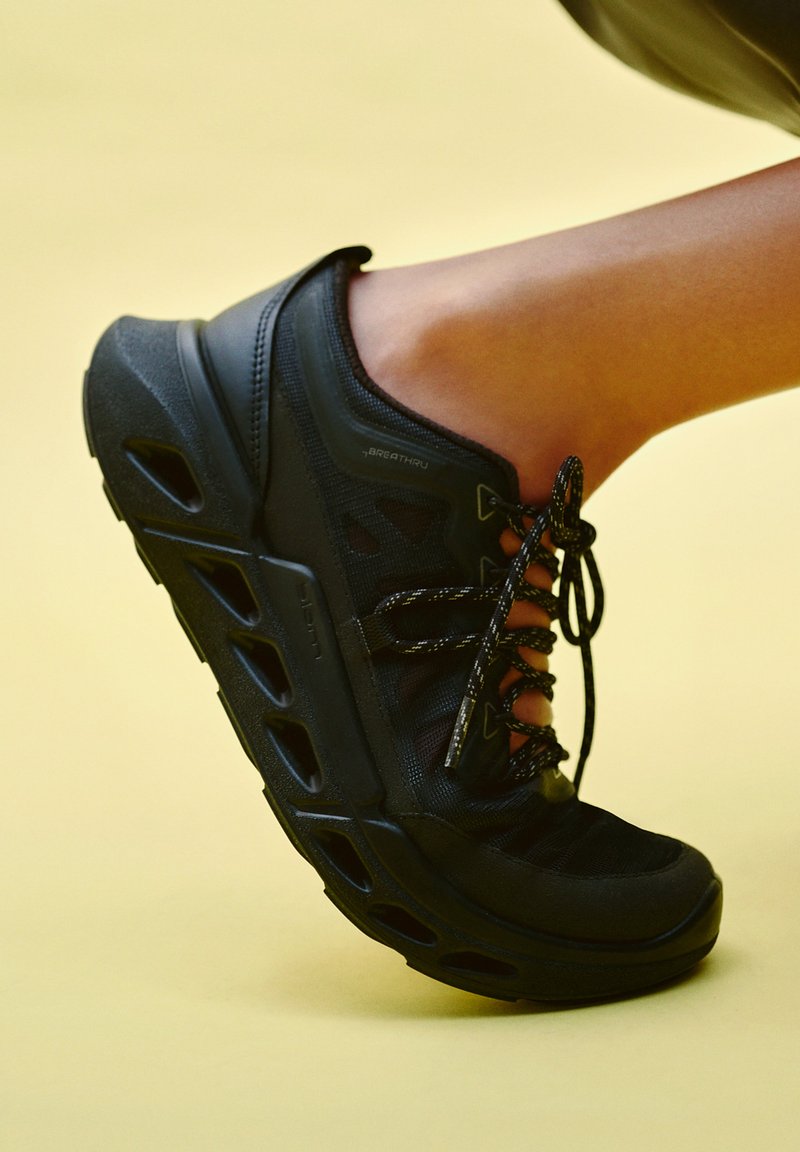 Musta urheilukenkä paksulla pohjalla ja avoimilla geometrisillä leikkauksilla, jalassa kohotetulla jalalla keltaista taustaa vasten.