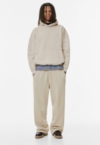 Beige, oversize kapucnis pulóver, elülső zsebfelülettel, párosítva a megfelelő melegítőnadrággal. Az anyag lágy és laza kinézetű, finom logórészlettel.