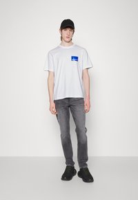 Balta T-kreklā ar zilu logo uz krūtīm, pelēkās džinsi un melnas sporta kurpes ar neona zaļiem akcentiem; modelis valkā melnu cepuri.