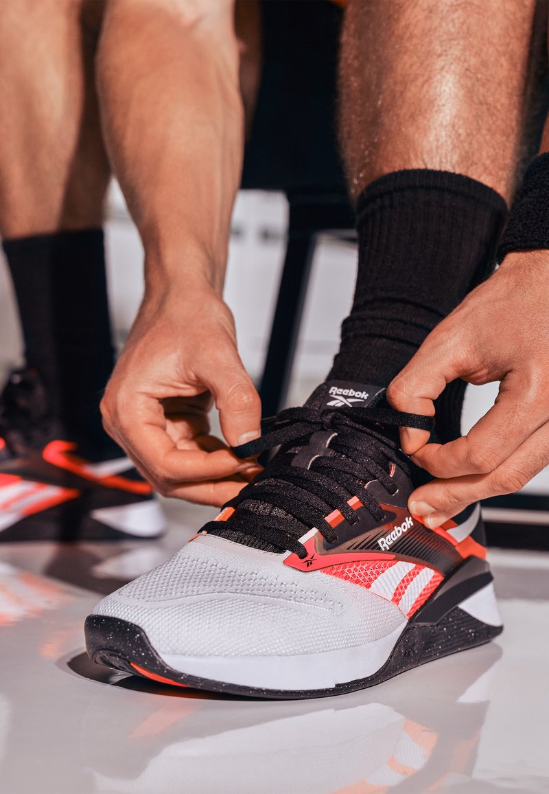 Persona atando los cordones de unas zapatillas Reebok negras y rojas, con calcetines negros, sentado sobre un suelo blanco y reflectante.
