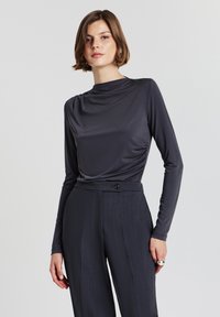 Maglietta a maniche lunghe, grigia scura, con scollo a barca e texture morbida e liscia, abbinata a pantaloni grigi scuri a vita alta e su misura, dotati di bottone.