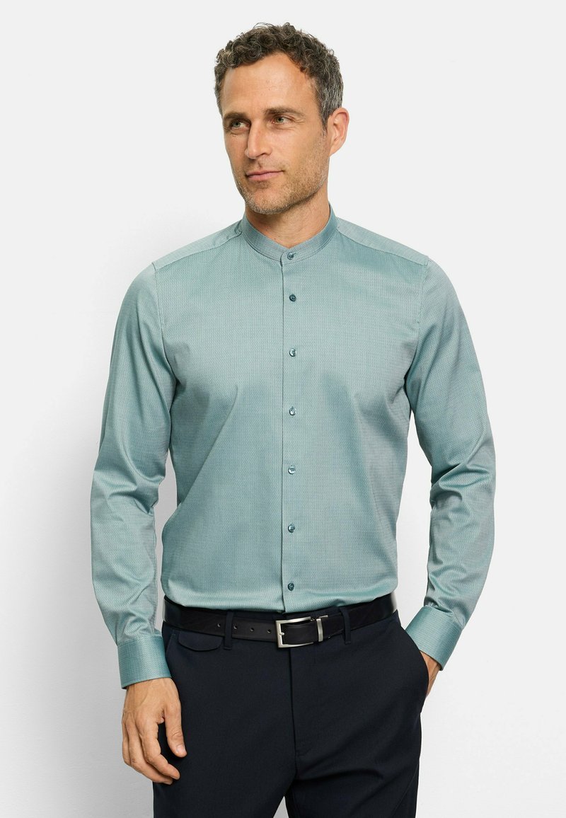 Langarmhemd aus türkisfarbenem Stoff mit einem subtilen Muster, Stehkragen und Knöpfen. Getragen mit dunklen Hosen und einem schwarzen Gürtel.