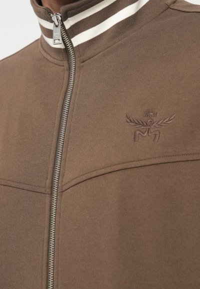 Casaco castanho com fecho éclair e colarinho alto, apresentando riscas brancas e castanhas, logótipo bordado e tecido texturado. Detalhe do fecho éclair prateado visível.