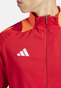 Rote Sportjacke mit orangefarbenen Akzenten, Reißverschluss, hohem Kragen und weißem Logo auf der Brust. Glatter Stoff.