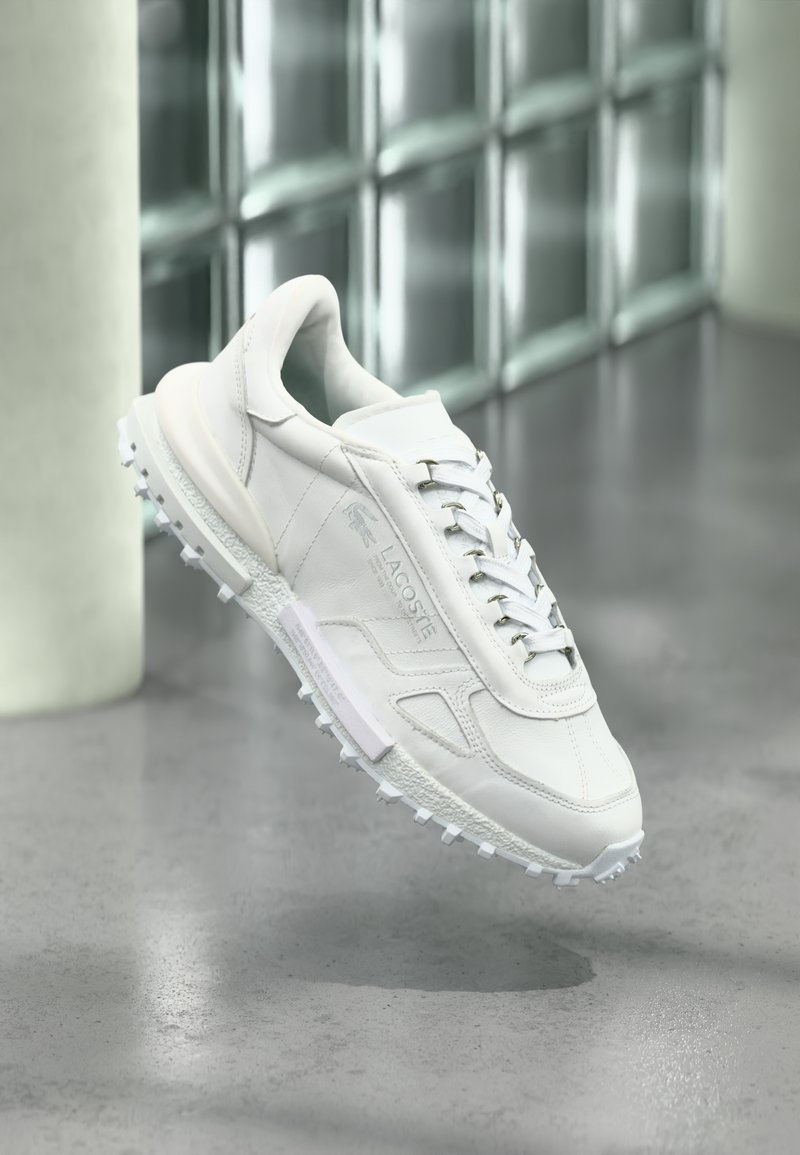 Chaussure de sport blanche en cuir perforé, avec des accents texturés et une semelle extérieure en caoutchouc antidérapante. Comprend des lacets argentés et un logo sur le côté.