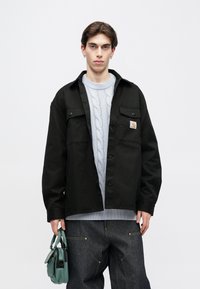 Czarna kurtka zapinana na guziki z dwoma kieszeniami na piersi, pod spodem szary sweter w warkocze, ciemne dżinsy oraz mała zielona torebka.