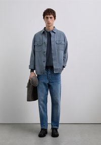 Jasnoniebieska kurtka dżinsowa z dwoma kieszeniami na piersiach, pod spodem granatowy sweter, niebieskie dżinsy i czarne buty. Trzyma szary wełniany kapelusz.