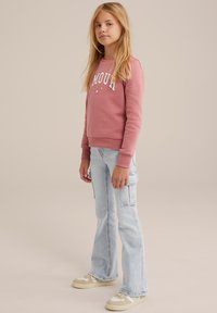 Růžová mikina s bílým textem "AMOUR" a hvězdami, zkombinovaná se světle modrými rozšířenými džíny s cargo kapsami a béžovými teniskami.