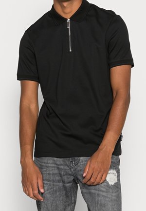 Czarna koszulka polo z zamkiem przy kołnierzu, krótkimi rękawami i kontrastowym wykończeniem, zestawiona z szarymi dżinsami z efektem postarzania. Gładka tekstura tkaniny.