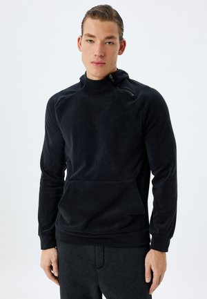 Sudadera de forro polar negro con collar alto, bolsillo frontal y detalles de cremallera. Textura suave y diseño ajustado, combinada con pantalones de chándal oscuros a juego.