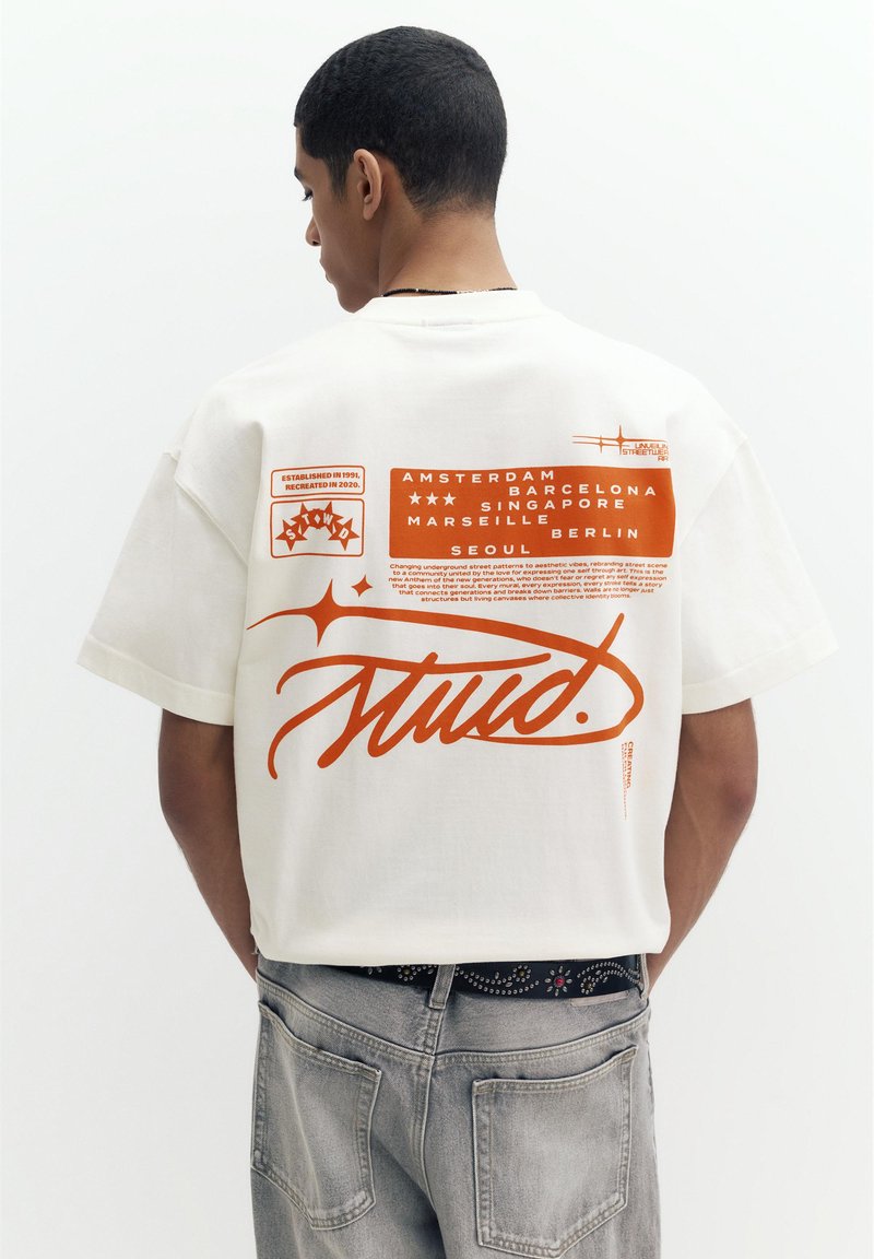 Ung mand set bagfra iført hvid t-shirt med orange grafisk tekst, der lister byer og stiliseret "Stüd" logo, kombineret med grå jeans.
