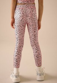 Leggings roses avec des taches de léopard noires, dotés de bandes latérales rouges, fabriqués dans un matériau extensible. Associés à des baskets blanches chunky.