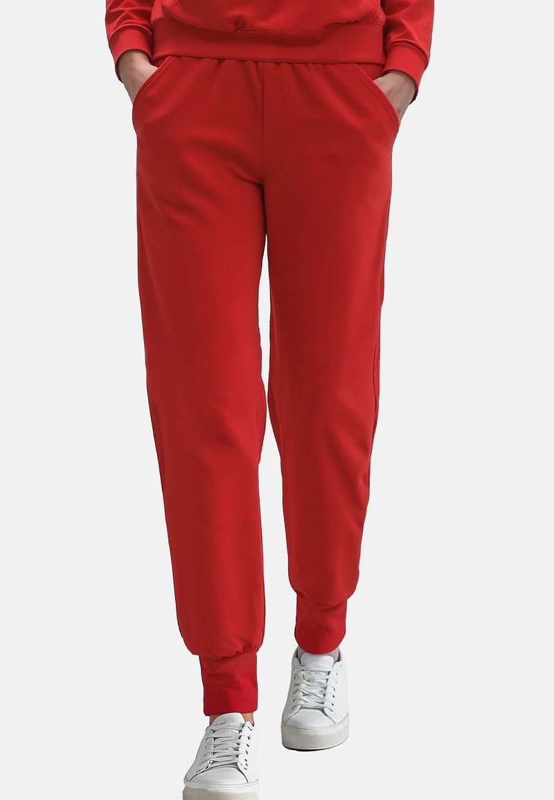 Røde sweatpants lavet af blødt stof, med elastisk talje, sidelommer og ribkanter ved anklerne, parret med hvide sneakers.