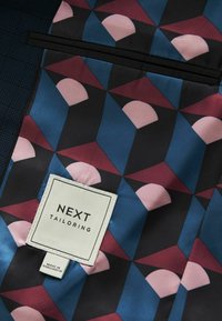 Kleurrijke geometrische voering met blauwe, roze en bordeauxrode patronen. Bevat een wit etiket met "NEXT TAILORING" en verzorgingsdetails.