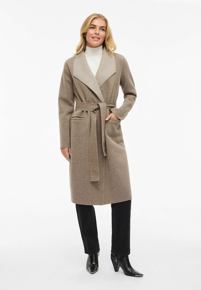 Cappotto lungo color marrone chiaro con un grande colletto, cintura e tasche, realizzato in tessuto morbido; abbinato a pantaloni neri e stivali neri con tacco.