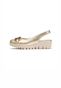 Zapato de tacón sintético dorado con puntera redondeada y una hebilla circular decorativa. Presenta una suela gruesa beige con bordes aserrados.
