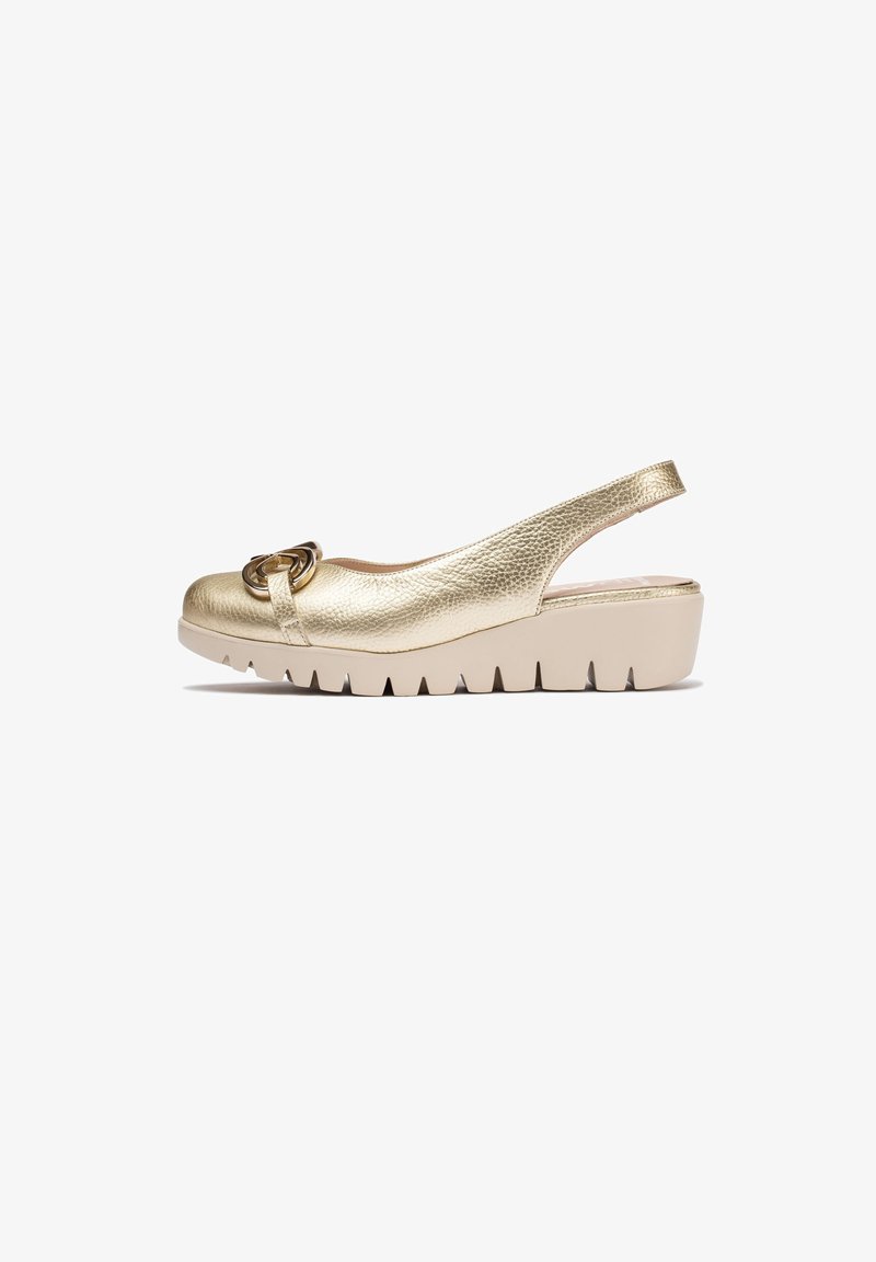 Zapato de tacón sintético dorado con puntera redondeada y una hebilla circular decorativa. Presenta una suela gruesa beige con bordes aserrados.
