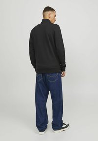 Sweat à capuche noir avec un col montant, associé à un jean en denim bleu ample. Détail notable : surpiqûres visibles des poches à l'arrière du jean.