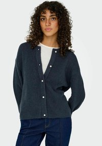Cardigan en tricot gris foncé à coupe lâche, avec un col en V et des boutons en perles. Porté par-dessus un top blanc et associé à un jean noir.