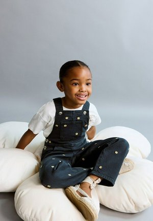 Fille souriante en salopette sombre à motifs floraux assise en tailleur sur un sol blanc rembourré, devant un fond gris.