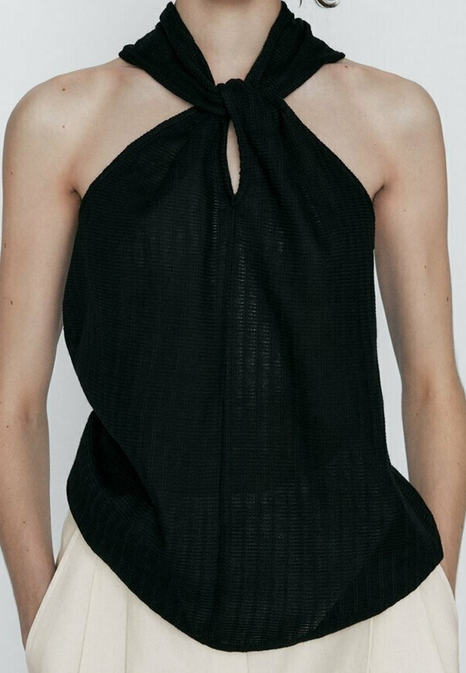 Femme portant un haut noir sans manches avec un nœud torsadé au cou et un pantalon beige sur un fond clair.