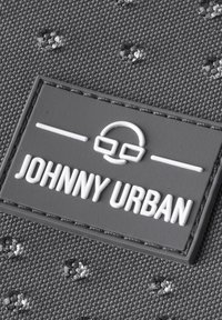 Taflă textilă neagră cu textură, având puncte ridicate, cu o etichetă rectangulare din cauciuc gri, gravată cu "JOHNNY URBAN" și un grafic cu ochelari.