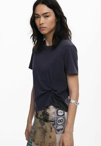 Dunkelgraues Baumwoll-T-Shirt mit verdrehtem Front-Detail, mit kurzen Ärmeln und lockerer Passform, kombiniert mit einem bedruckten Rock.