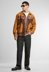 Braun- und beige-farbige, blockierte Jacke mit großen Taschen, Knopfverschluss und lederähnlicher Textur, kombiniert mit dunklen Jeans und schwarzen Schuhen.