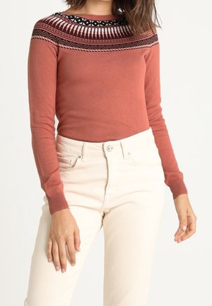 Maglione lavorato a maglia di colore ruggine con un design a motivi in bianco e nero attorno all'collo. Abbinato a jeans aderenti di colore chiaro.