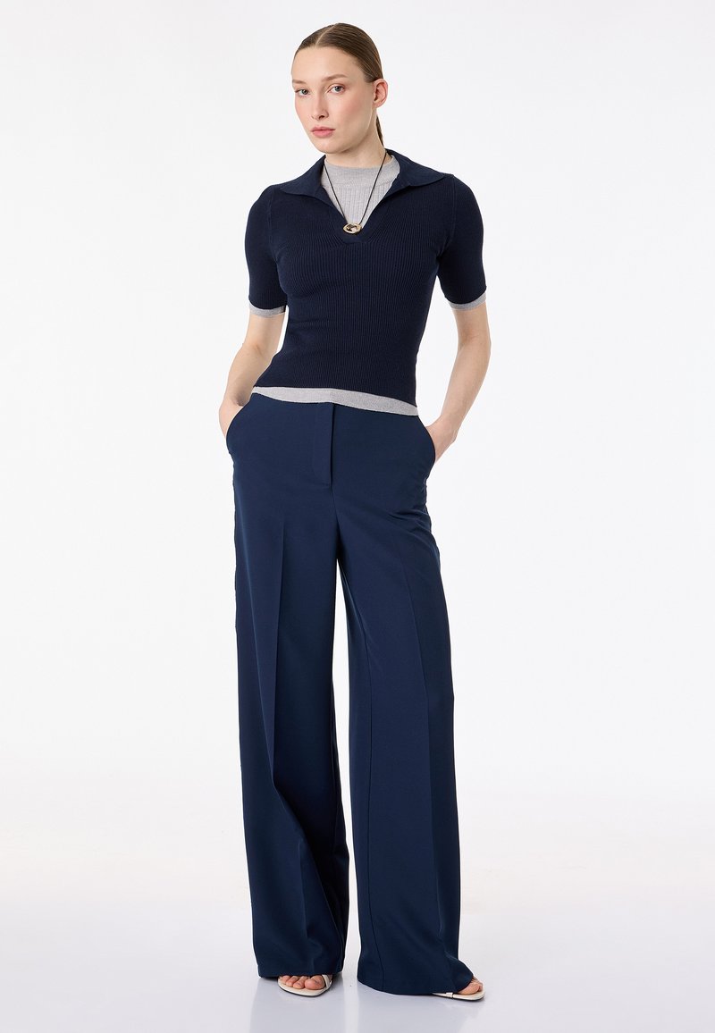 Vrouw staat met handen in de zakken, draagt een marineblauw shirt met korte mouwen over een grijs shirt, wijd uitlopende marineblauwe pantalon en sandalen.