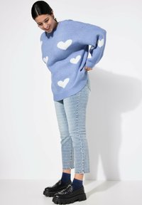 Světle modrý oversized svetr s bílými vzory srdcí, doplněný o strukturované světle modré džíny a černé platformové mokasíny s hroty.