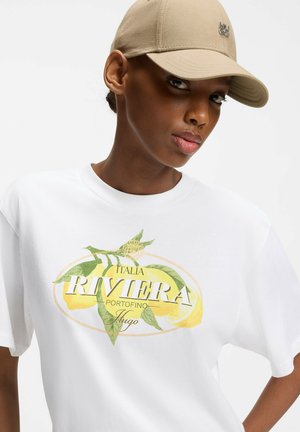 Frau trägt beige Kappe und weißes T-Shirt mit gelbem Zitronen- und grünem Blätterdesign mit der Aufschrift „Italia Riviera Portofino Hugo“ vorne.