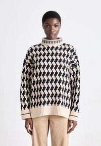 Strickpullover in Beige und Schwarz mit einem Kreuzmuster, ausgestattet mit einem hohen Kragen und gerippten Säumen. Getragen mit beigen Hosen.