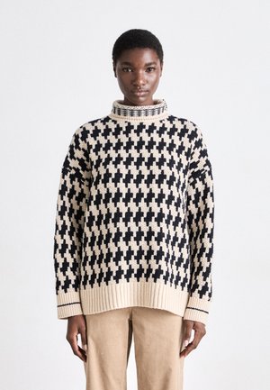 Strickpullover in Beige und Schwarz mit einem Kreuzmuster, ausgestattet mit einem hohen Kragen und gerippten Säumen. Getragen mit beigen Hosen.