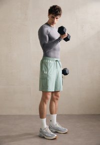 Pilkos spalvos ilgomis rankovėmis sportinė maikutė, šviesiai žali šortai su raišteliu ir šoninėmis kišenėmis, pilki ir balti sportbačiai, laiko juodus hantelius.