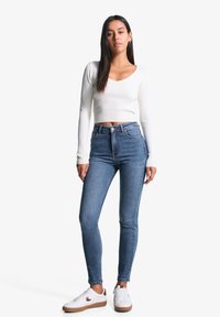 Sinisiä korkeavyötäröisiä skinny-farkkuja, jotka on valmistettu denimistä, yhdistetty valkoiseen ribatusta pitkähihasta crop-topiin ja valkoisiin tennareihin, joissa on ruskeita yksityiskohtia.