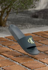 Sandale glissante grise avec un design texturé. Présente une large bride avec un logo "CK" jaune. Semelle intérieure lisse et semelle en caoutchouc sur une surface en briques.