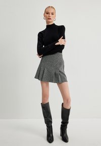 Suéter negro de cuello alto, falda plisada gris con estampado y botas de tacón alto negras hasta la rodilla. La modelo está de pie contra un fondo liso.