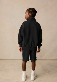 Svart bomullsoverdel med hög krage i oversized stil, parat med svarta shorts. Modellen bär vita strumpor och sneakers, står på en neutral bakgrund.