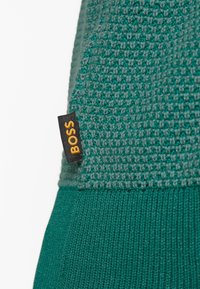 Gros plan sur un tissu tricoté vert avec un motif texturé et une petite étiquette noire portant l'inscription « BOSS » en lettres jaunes.