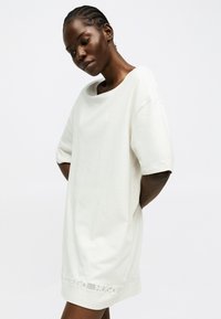 Γυναίκα με πλεξούδες, φοράει ένα υπερμεγέθες λευκό φόρεμα με κοντά μανίκια και το κείμενο «HUGO» κοντά στο τελείωμα, στέκεται μπροστά σε απλό φόντο.