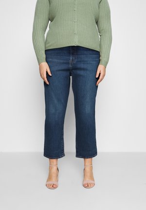 Kvinde iført mørkeblå korte jeans, lysegrøn ribstrikket trøje med knapper og beige åbne hæle, stående på en ensfarvet baggrund.