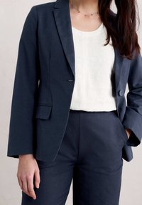 Navy blue tailored blazer with a single button, paired with a white top and matching trousers. Smooth texture, structured shoulders, minimal design.