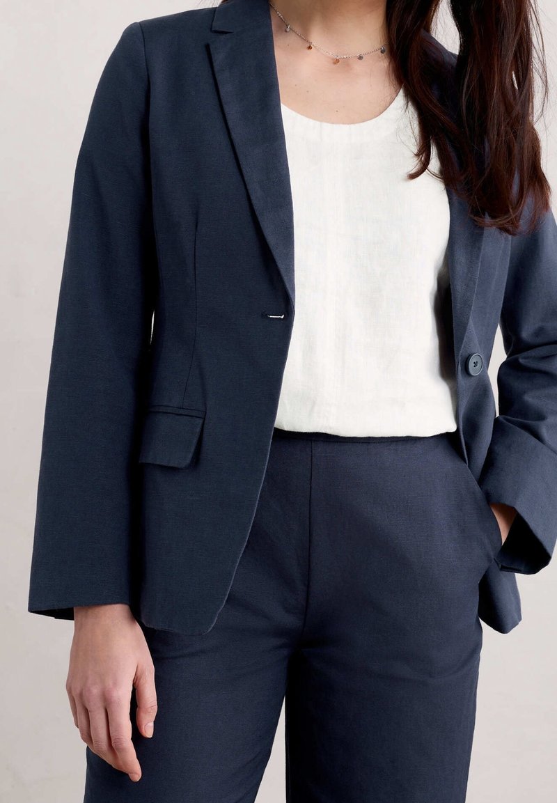 Navy blue tailored blazer with a single button, paired with a white top and matching trousers. Smooth texture, structured shoulders, minimal design.