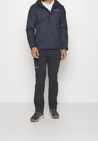 Marineblaue wasserdichte Jacke mit Kapuze und Reißverschlusstasche; kombiniert mit schwarzen Hosen und grauen Mesh-Sneakern. Schlichte, funktionale Outdoor-Bekleidung.