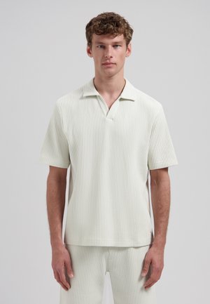 Man draagt een korte mouwen, gestructureerd, gebroken wit poloshirt en bijpassende gestructureerde broek, staand tegen een effen lichtgrijze achtergrond.