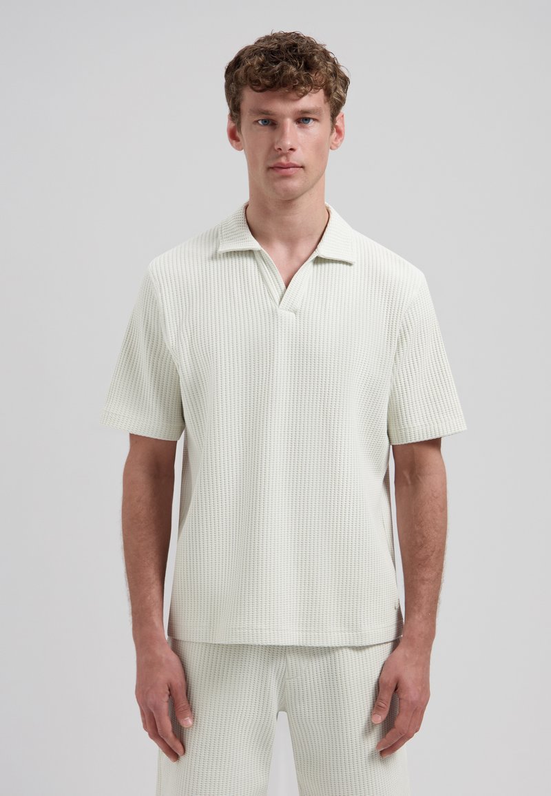 Homme portant un polo à manches courtes, texturé, de couleur blanc cassé, et un pantalon texturé assorti, debout devant un fond uni gris clair.