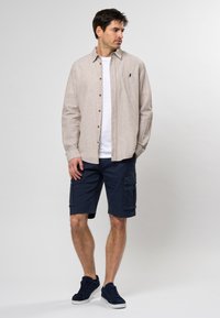 Camisa de lino beige de botones sobre una camiseta blanca, combinada con pantalones cortos de carga azul marino. El conjunto incluye zapatillas de ante azul marino y una silueta estructurada.