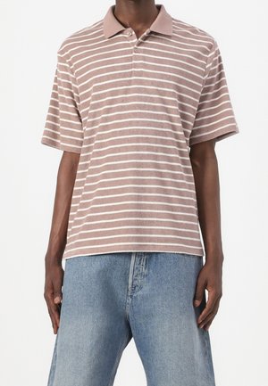 Personne portant un polo à manches courtes marron clair avec des rayures horizontales blanches et un jean en denim bleu clair.